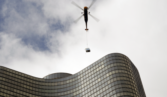 A heavy-duty helicopter delivering another high-efficiency commercial boiler to the rooftop of Lake Point Tower against its iconic curved glass facade during a massive Chicago HVAC upgrade.