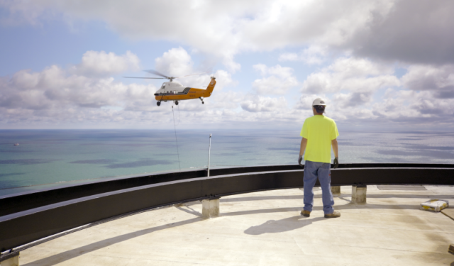A commercial HVAC technician on the roof of Lake Point Tower observes a heavy-duty helicopter performing a massive high-rise boiler lift against the backdrop of Lake Michigan.