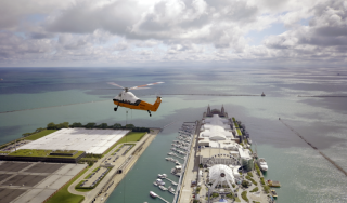 Chicago Commercial HVAC - Aerial Helicopter Rigging Mid-Flight A stunning aerial view of a heavy-duty helicopter ascending with a commercial boiler during a complex high-rise HVAC lift at Lake Point Tower, with Navy Pier and Lake Michigan in the background.