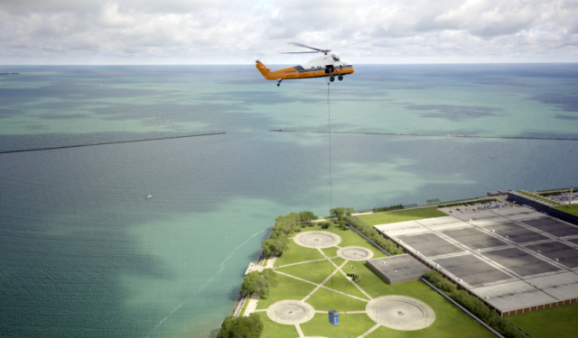 A heavy-duty helicopter lifting a massive commercial boiler over a Chicago memorial park during a complex high-rise HVAC rigging operation at Lake Point Tower.