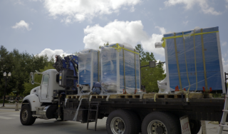 Chicago Commercial HVAC - Boiler Transport & Staging Logistics Side profile of a flatbed truck transporting four massive, shrink-wrapped commercial boilers to stage for a complex high-rise HVAC helicopter lift at Lake Point Tower in Chicago.