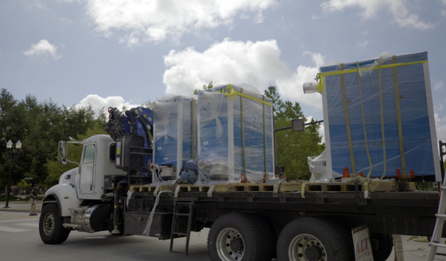 Side profile of a flatbed truck transporting four massive, shrink-wrapped commercial boilers to stage for a complex high-rise HVAC helicopter lift at Lake Point Tower in Chicago.